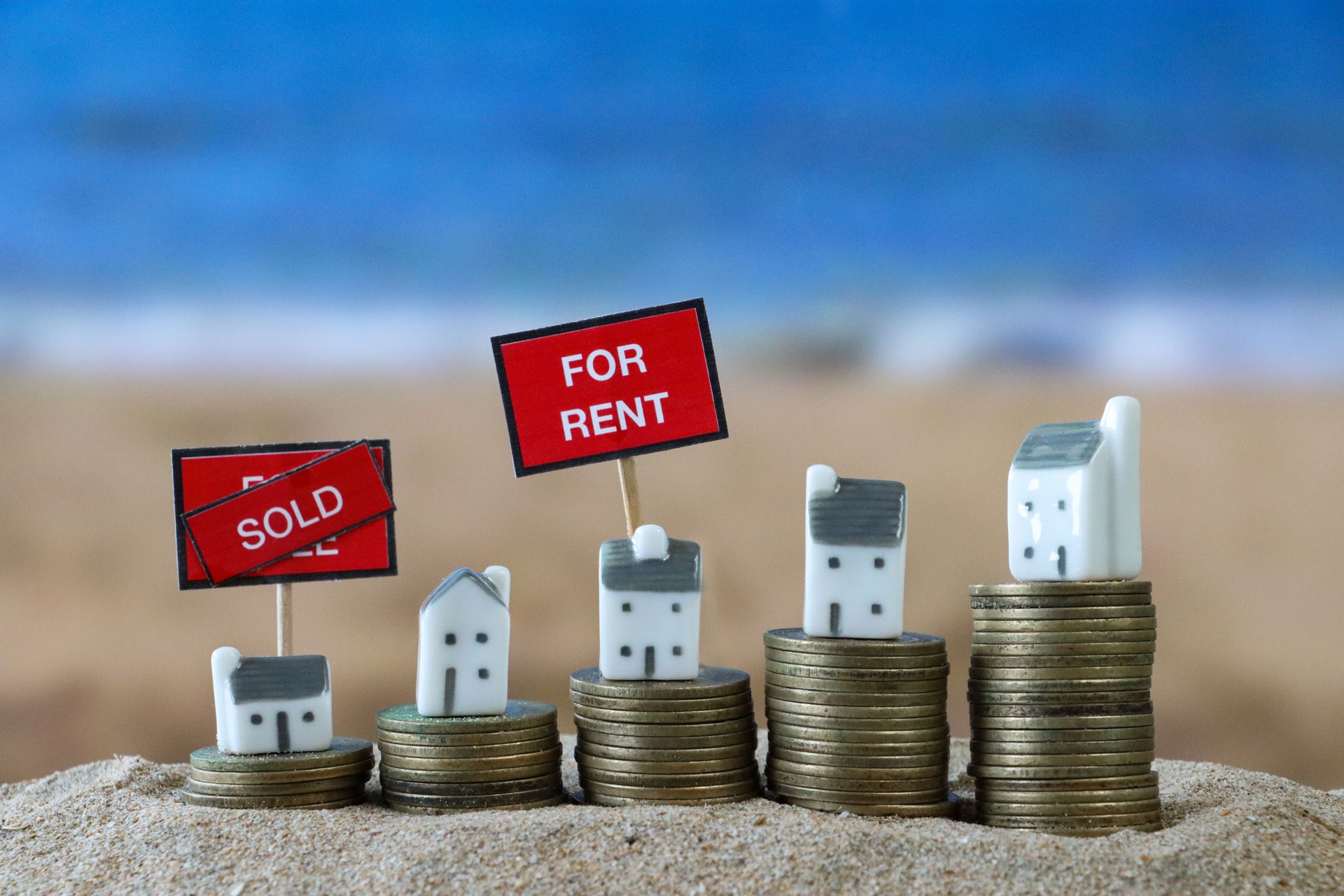 Close-up image of 'For Rent' sign beside row of model holiday cottages stood on piles of coins, sandy beach background with waves breaking at low tide, sea and coastline, holiday rental and vacation home concept, focus on foreground